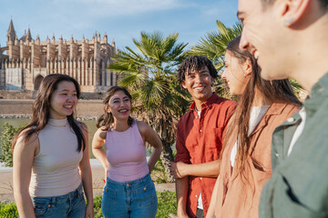 Young multicultural friends talking and smiling in Mallorca - Group of diverse young people enjoying a conversation outdoors near cathedral. Concept of travel, friendship and youth.