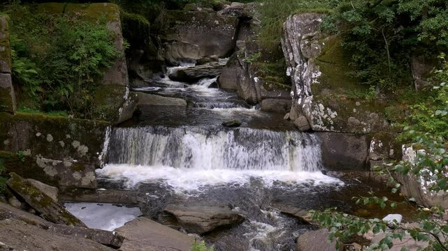 Fast-flowing waterfalls cascade over rocks in a lush, green landscape in Scotland. A dynamic and scenic natural environment.