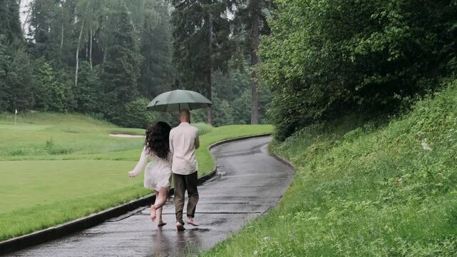 A young couple in love runs along a path with an umbrella on a rainy summer day