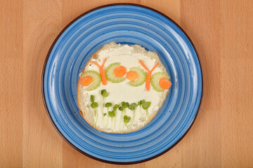 A creatively decorated open-faced sandwich, featuring cream cheese, sprout "grass," and vegetable "butterflies" made from celery and carrot, rests on a blue plate atop a wooden surface