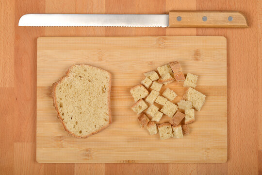 A single slice of bread sits beside a pile of irregularly cut bread cubes on a light wooden cutting board, accompanied by a bread knife