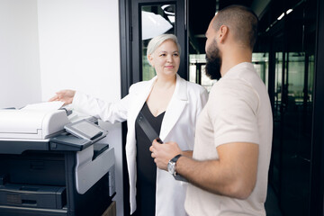 Obraz premium Two colleagues engaged in conversation beside printer in modern office. one woman in white coat with short grey hair, and man with beard in casual attire.