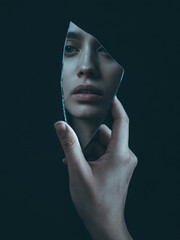 A dramatic, close-up portrait photograph captures a hand delicately holding a shard of a shattered mirror, reflecting a distorted image of a woman’s face