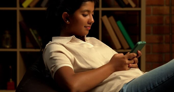 Indian Teenage Girl Using Smartphone While Sitting Comfortably on a Bean Bag at Home with Moody Lighting Against Brick Wall, Scrolling or Staying Connected with Friends Through Social Media
