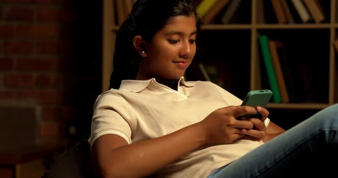 Indian Teenage Girl Using Smartphone While Sitting Comfortably on a Bean Bag at Home with Moody Lighting Against Brick Wall, Scrolling or Staying Connected with Friends Through Social Media