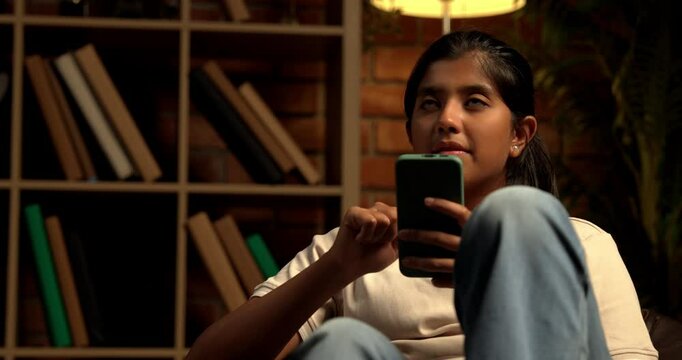 Indian Teenage Girl Using Smartphone While Sitting Comfortably on a Bean Bag at Home with Moody Lighting Against Brick Wall, Scrolling or Staying Connected with Friends Through Social Media