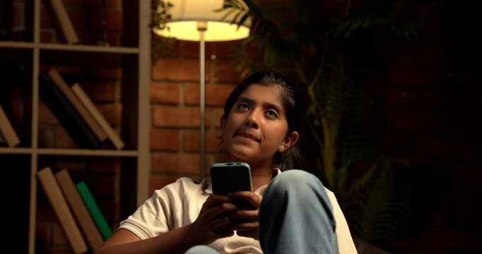Indian Teenage Girl Using Smartphone While Sitting Comfortably on a Bean Bag at Home with Moody Lighting Against Brick Wall, Scrolling or Staying Connected with Friends Through Social Media