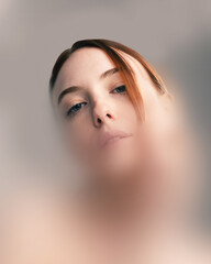 Conceptual artwork. Portrait of red-haired woman with piercing blue eyes looking downwards through blurred filter, conveying isolation and introspection. Concept of beauty, psychology.