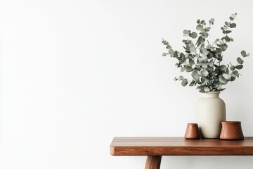 Simple, minimalist scene.  A vase of eucalyptus sits on a light-colored wooden table against a white wall