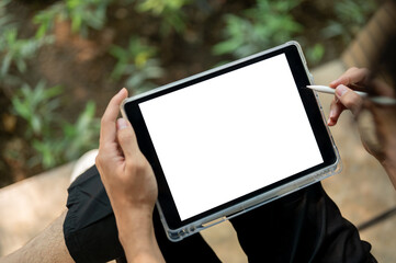 Close up of a hand holding white screen tablet and stylus while sitting cross legged near plant bed.