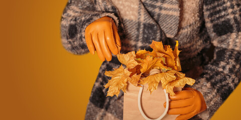 Woman in coat and yellow gloves holding kraft paper shopping bag full of autumn dried leaves. Autumn background