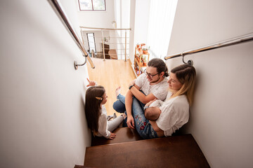 Young family with newborn baby and daughter are sitting on stairs in their modern home, talking and...