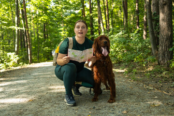 A cheerful woman kneels beside her Irish Setter dog while holding a map and pointing ahead on a...