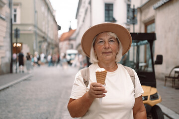 60s senior woman walking leisurely down a city street with a cone of ice cream, enjoying a moment...