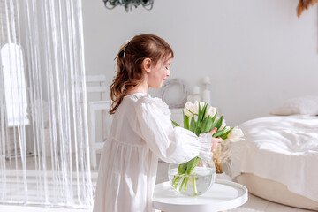 Cute little girl in a white dress arranging a bouquet of white tulips in a glass vase on a white...