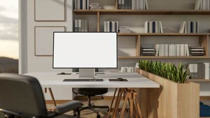 Blank screen computer on wooden table with swivel chair in office with large bookshelf on white wall