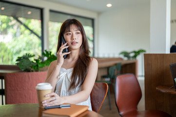 Asian woman in camisole talking on the phone and grabbing coffee while sitting at cafe wooden table.