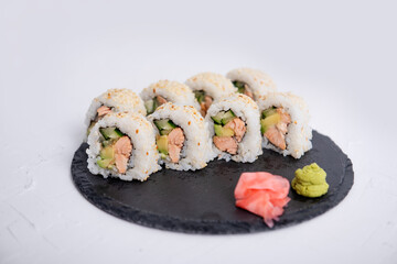 Close-up of sushi California rolls with salmon, avocado, and cucumber, topped with sesame seeds. Served with wasabi and pickled ginger on a black slate plate.