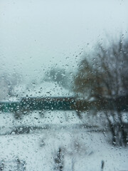 Water drops condensation on window glass. View of winter snowy street with houses. Selective focus.
