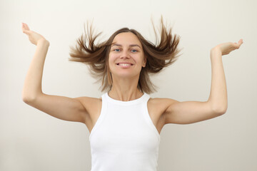 Happy young woman with flying hair smiling at camera on white backdrop. Casual natural look, raised arms, carefree emotion. Concept of freedom, fun, energy, youth branding, wellness, personal care