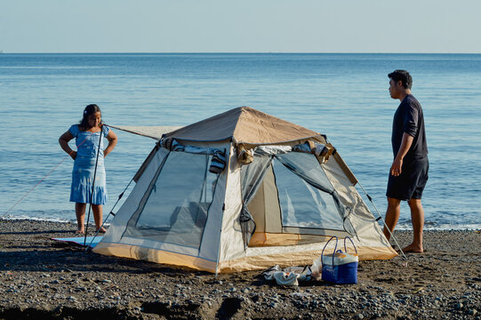 A father and his daughter carefully inspect their newly set-up beach tent, preparing for a fun day.
