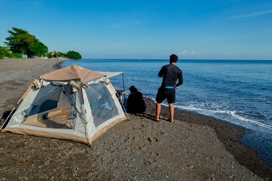 A father stands while other family members remain seated in front of the tent on a coastal beach. - Powered by Adobe