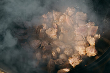 Pieces of meat frying in oil in a large cast iron cauldron with steam. Cooking outdoor in a big pot...