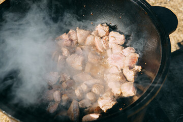 Pieces of meat frying in oil in a large cast iron cauldron with steam. Cooking outdoor in a big pot...