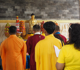 Fototapeta premium A group of individuals in yellow and orange robes stand facing a shrine featuring a Buddha statue. Offerings of fruit are stacked around the statue. The setting suggests a Buddhist temple with a stone