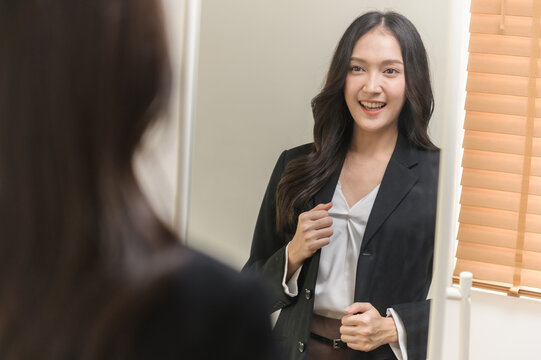 Young beautiful Asian woman in formal suit checking dressing tidiness and encourage herself in mirror at home before go for job interview. Woman recruitment employee or staff in company concept.