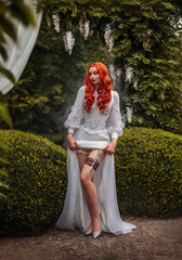 Fantasy red-haired dangerous woman medieval assassin queen with dagger in sheath on leg leather belt. Green vintage old style dress. Girl princess warrior. Garden summer nature trees. Red long hair