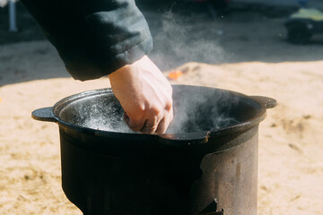 Person cooking in a cauldron with steam rising. Outdoor food preparation in a cast iron pot on a...