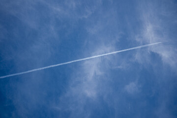 An airplane contrail streaks across a blue sky with wispy clouds.