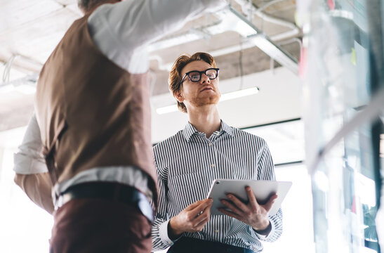 Young Caucasian businessman holds tablet while listening to older colleague explaining project on glass board in bright office. They stand near sticky notes and charts during planning discussion