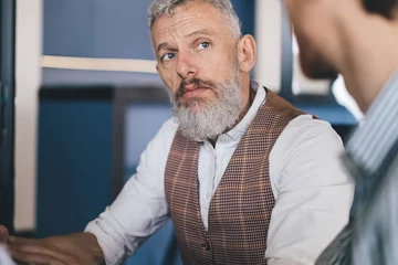 Gordijnen Close-up of senior white businessman with gray hair and beard attentively listening during serious business discussion, dressed in classic office wear, seated in modern workplace with blue tones © BullRun