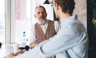 Obraz premium Cheerful young Caucasian businessman in glasses smiles and gestures toward the camera in modern office, while older colleague in background laughs. Friendly team dynamic, casual environment