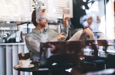 Young Caucasian businessman in striped shirt smiling and gesturing while discussing project with older colleague in vest. Casual business meeting in cozy modern café, natural daylight, spring mood