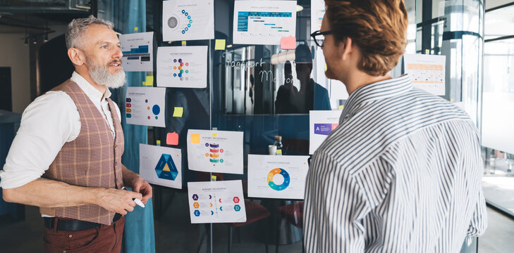 A senior White businessman explains business data to a younger colleague in front of a glass wall filled with colorful charts and infographics in a creative open-plan workspace