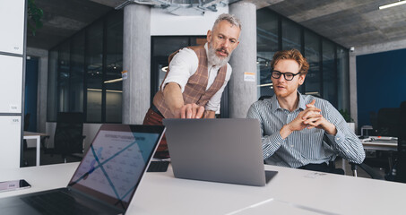Senior businessman guides young colleague by pointing at laptop screen during a mentoring session...