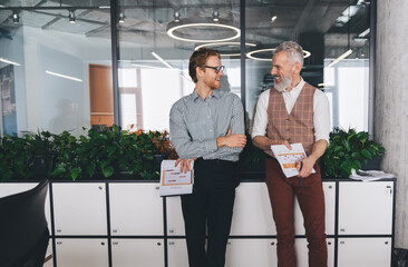Two Caucasian businessmen, one young and one older with a gray beard, stand by office lockers holding charts, smiling and chatting casually in a modern, creative workspace