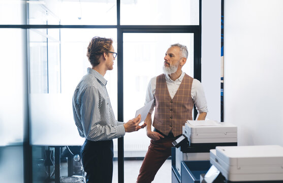 Two male colleagues, one senior and one younger, talk near an office printer in bright daylight, discussing document printing and planning during a casual business conversation