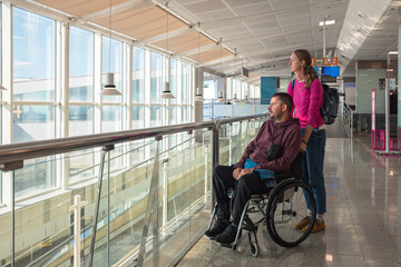 Man with disability using wheelchair with his female personal assistant is at the airport, looking...