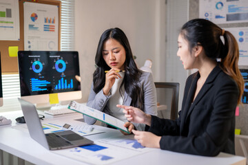 Two women are sitting at a desk with a laptop and a stack of papers