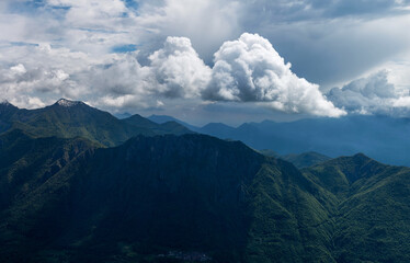 Atmospheric panorama of Pizzi di Parlasco, highlighting the beautiful interplay of light and shadow across verdant forested slopes, with dramatic clouds enveloping the peaks. Valsassina, Italy