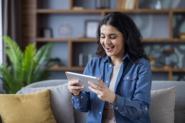 A smiling woman enjoys time on her tablet, relaxing on a comfortable sofa at home with a plant.