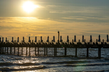 Silhouetted pier with closed umbrellas and loungers at sunset, ready for new tourist season. Sparkling sea under warm Mediterranean light. Side, Antalya, Turkey