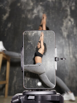 Fitness influencer doing home workout and stretching on yoga mat while live streaming on smartphone. Sporty young woman filming online fitness training for social media followers