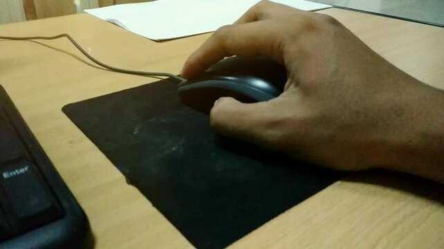 A person&rsquo;s hand using a wired computer mouse on a black mousepad, placed on a wooden desk next to a keyboard, in an office or home workspace.