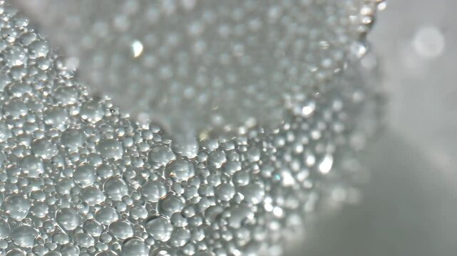 Close-up of transparent liquid foam with bubble clusters forming textured cube edges and surface reflections against light backdrop.