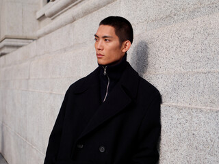 Obraz premium Portrait of handsome Chinese young man wearing black overcoat posing in the street, young guy with black short hair all in black with urban background. Male fashion, cool Asian young man lifestyle.